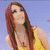 A woman with long red hair wears a yellow sleeveless top and pink leg warmers, seated on a yellow vehicle with a blue sky background.