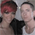 Two people stand close together; one has short dark hair with red highlights, the other has short dark hair, both wear dark and light tops against a light background.