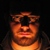 Pixelated static image of a person with dark hair and a dark top against a black background, wearing a brown headpiece with a central black rectangular section.