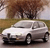 A silver sedan with black wheels is positioned on a road, with trees and a grassy field in the background.