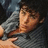 Pixelated close-up of a person with light skin wearing a gray shirt, head tilted slightly right, eyes closed, mouth closed, against dark background.