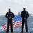 Two uniformed figures stand side-by-side against a blurred background with an American flag, overlaid with red text blocks.