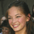 A pixelated close-up of a smiling person with dark hair tied back, showing a portion of the face and neck against a dark background.