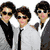 Three people in formal attire stand together on a white background, with one person wearing sunglasses and another holding a peace symbol drawn in sand.