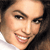 Pixelated close-up of a smiling person with light skin, dark hair, and visible teeth, wearing a white garment against a dark background.