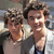 Two individuals with dark curly hair stand close together, one wearing a light-colored shirt and the other in a dark shirt with a white collar, against a blurred background.