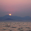 Sunset over water with a mountain silhouette and a bright yellow-orange sun partially visible above the peak.