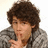 Pixelated image of a person with brown curly hair wearing a gray jacket, holding an object near their face against a light background.