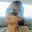 A person with dark hair tied back wears sunglasses and a light-colored top, seated in what appears to be a vehicle interior.