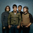 Four individuals stand in a against a blue background, with red pixelated text on dark red and gray