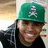 A person wearing a green cap with white skull and crossbones symbol, black hoodie, smiling face, on left side of image; right side has bright green pixelated text on black background.