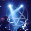 A concert scene with performers on stage under blue lighting, featuring a large inverted pentagram backdrop and audience silhouettes.