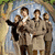 Group of five people in brown uniforms standing under an archway with greenery, one person raising a black arm.