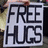 A static image with a bright green background, featuring a white sign with black text reading "FREE HUGS" and scattered blue pixelated text fragments.