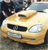 Pixelated image of a light-colored car with dark windows and headlights, positioned on a textured surface under a blurred background.