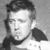 Black-and-white pixelated portrait of a person with short dark hair, closed eyes, and a neutral expression wearing a dark collar or neckband.