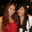 Two people with long brown hair stand close together, one in a red sleeveless top and the other in dark clothing with white accents against a dark background.