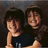 Two pixelated figures with dark hair and light skin pose closely together against a dark background, one figure holding the other's shoulder.