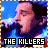 Pixelated image of a person singing into a microphone with blue lighting and text reading THE KILLERS at the bottom, framed by a yellow border.