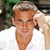 Pixelated static image of a person with light brown hair wearing a white shirt, hand raised near head, against blurred green and dark background.