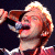 Pixelated image of a person with light-colored hair holding a microphone close to their mouth, wearing a dark top with a light-colored collar.