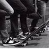 Black-and-white image of three people's lower bodies standing on a skateboard, with visible shoes and pants.