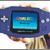 A split-screen image showing a blue Game Boy Advance device held in hands on the left and green pixelated text "Original Icons .com" on a dark background on the right, with visible pixelation and dithering effects.
