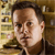 Static pixelated image of a person with short dark hair, light skin, and a neutral expression, set against a blurred background with indistinct shapes and warm tones.