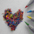 A heart-shaped icon made of colorful pixel clusters on a light gray background with scattered crayons.