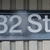 A static image of a street sign displaying '32 St' in white text on a blue background with a striped pattern.