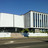 Pixelated static image of a modern building with white and gray sections under a clear blue sky, featuring a dark horizontal band and greenery below.