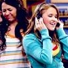 Two women with long hair, one in a striped top and the other in a teal sweater, both holding phones to their ears in a brightly lit indoor setting.