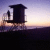 Pixelated silhouette of a dark structure against a gradient sky transitioning from purple to orange, with horizontal bands of color and blurred background elements.