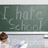 A child with brown hair in a bun writes on a green chalkboard that reads "I hate School" while seated at a desk.