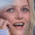 Pixelated close-up of a person with light skin tone, blue eyes, and light brown hair, mouth open showing teeth, wearing a light blue checkered garment.