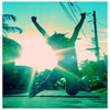 Silhouetted figure with arms raised and legs spread in mid-air against a bright sunburst, green-tinted background with trees and utility poles.