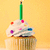 Pixelated cupcake with white frosting, colorful sprinkles, and a green candle on top against a yellow background.
