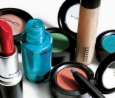 Static close-up of various makeup products including lipstick, eyeshadow compacts, and a bottle on a white surface with black circular borders.