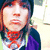 A person with dark hair and a beanie wears a purple jacket over a white shirt, red patterned scarf, and green backpack straps; the background is blurred outdoor scenery.