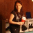 A pixelated image of a person with long dark hair holding a stemmed glass containing red liquid, seated at a table in front of a brown background.