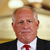 Static pixelated image of a person in formal attire with light skin, short white hair, wearing a black suit jacket, white dress shirt, and red tie against a blurred golden background.