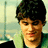 Pixelated portrait of a person with dark hair and green clothing against a blurred background with blue text reading 'John Maye' on the right.