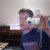 Pixelated image of a person with short dark hair wearing a dark blue shirt, holding a white rectangular object near their face against a light background.