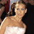 A pixelated image of a person with light brown hair styled in a side ponytail wearing a strapless white dress against a dark background with blurred figures.