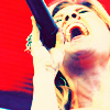 A person with light-colored hair sings into a black microphone against a red background, mouth open wide in a close-up shot.