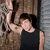 A person in a black sleeveless shirt and dark cap poses with one arm raised, against a textured background.