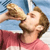 A person with light brown hair and a beard wears a red shirt, drinking from a bottle with a blue background.