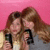 Two figures with long brown hair embrace against a solid pink background, one holding a black smartphone-like device.