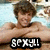 A shirtless person with brown hair lies in a pool, arms crossed over chest, smiling at the camera.