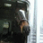 Pixelated static image of a figure in dark clothing holding an object near their face against a light background.